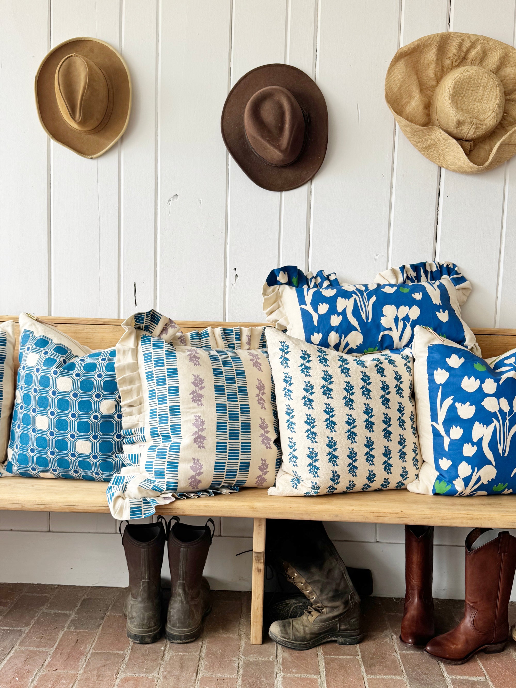 pillows gathered on wood bench with hats hanging on white wall and farm boots beneath bench